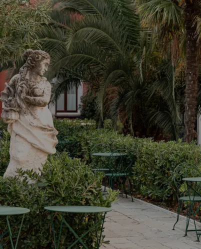Outdoor garden with a stone statue of a woman carrying a basket, surrounded by green bushes and metal chairs and tables.