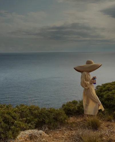 Person wearing a large sunhat and light dress standing on a shrub-covered cliff overlooking a calm sea under a cloudy sky.