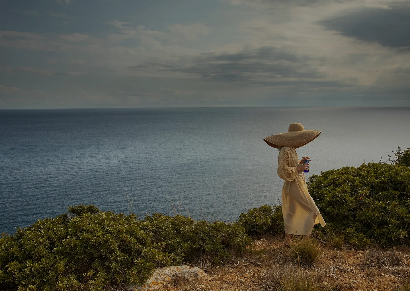 Woman wearing a large wide-brimmed hat and light dress standing on a cliff overlooking a calm sea under a cloudy sky.