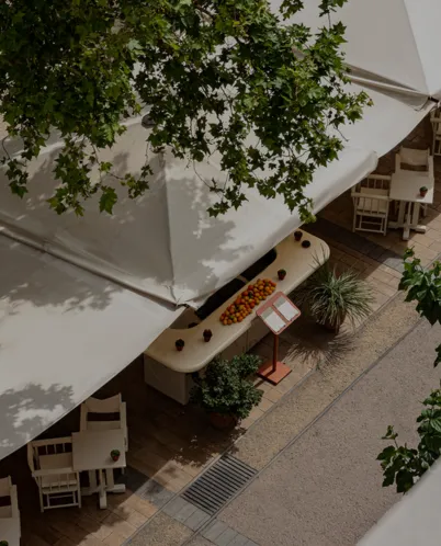 Overhead view of an outdoor café with white umbrellas, wooden tables, chairs, a long fruit display table, and surrounding green foliage.
