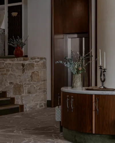 Elegant hotel lobby corner with stone wall, wooden furniture, floral arrangement in a vase, and a two-candle holder on a marble top.