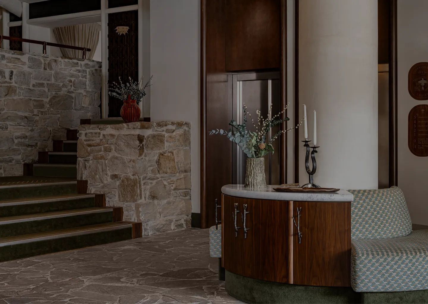 Elegant hotel lobby with curved wooden seating, stone flooring, stone walls, and decorative floral arrangements and candlesticks.