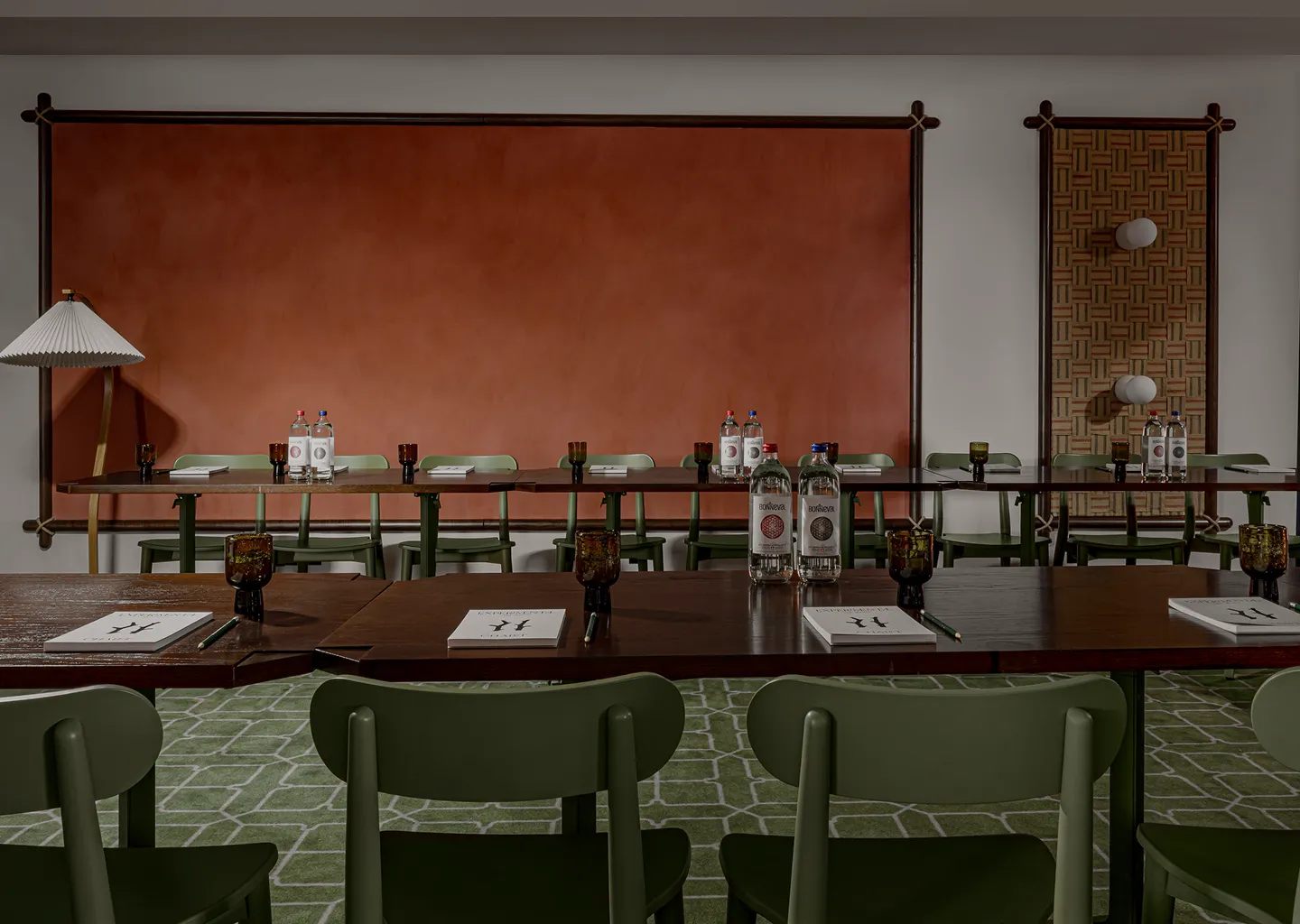 Conference room with dark wooden tables arranged in rows, green chairs, notebooks, pencils, and bottles of water against a terracotta wall and patterned wall panel.