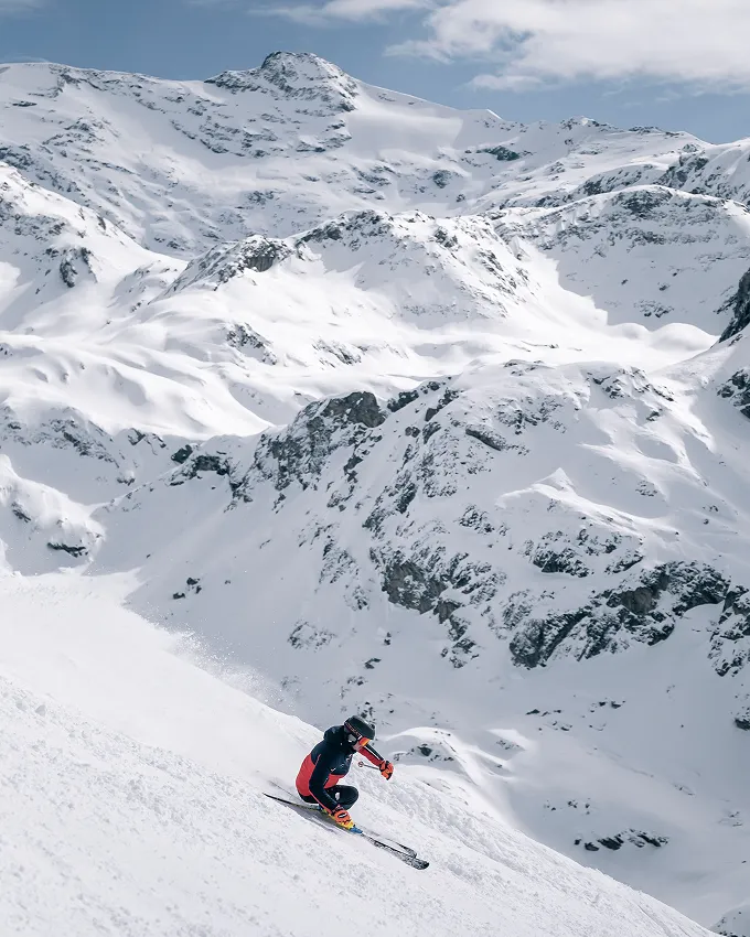 Skier in black and red outfit skiing downhill on a snowy mountain with snow-covered peaks in the background.