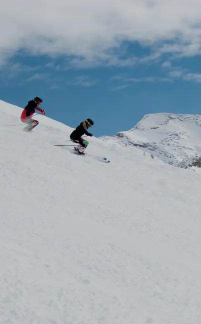 Two skiers descending a snowy mountain slope under a partly cloudy blue sky.
