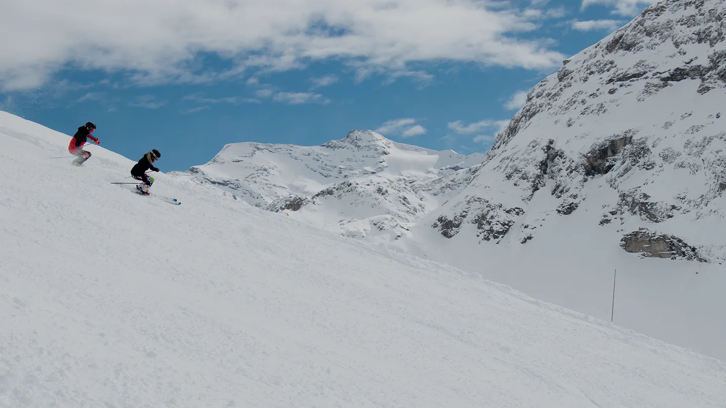 Two skiers descending a snowy slope with snow-covered mountains and a partly cloudy sky in the background.