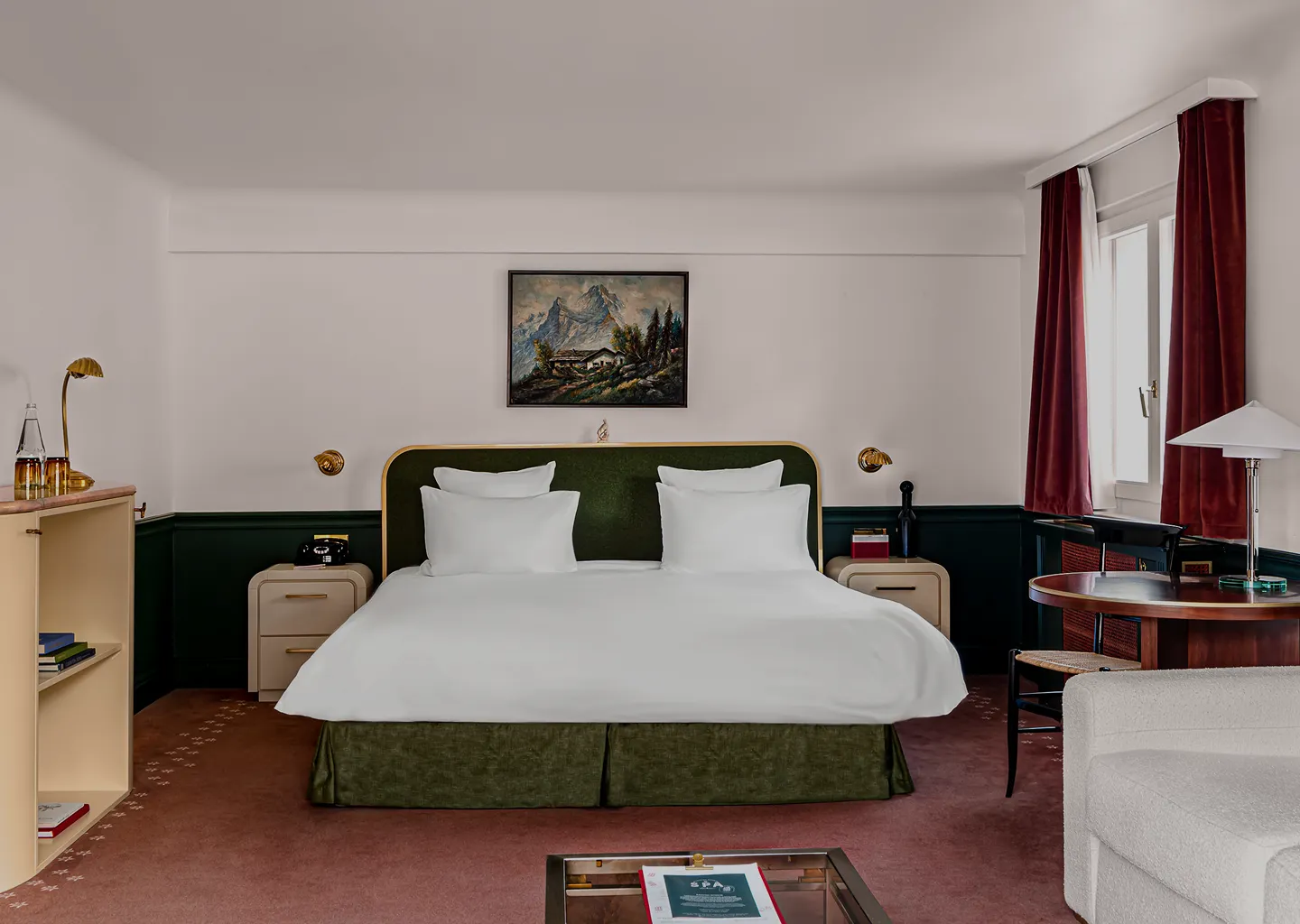 Modern hotel room with a large bed, green upholstered headboard, white bedding, red curtains, and a round wooden table.