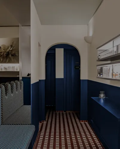 Interior hallway with navy blue lower walls, white upper walls, patterned floor tiles, patterned upholstered bench on the left, and glass shelves on the right.