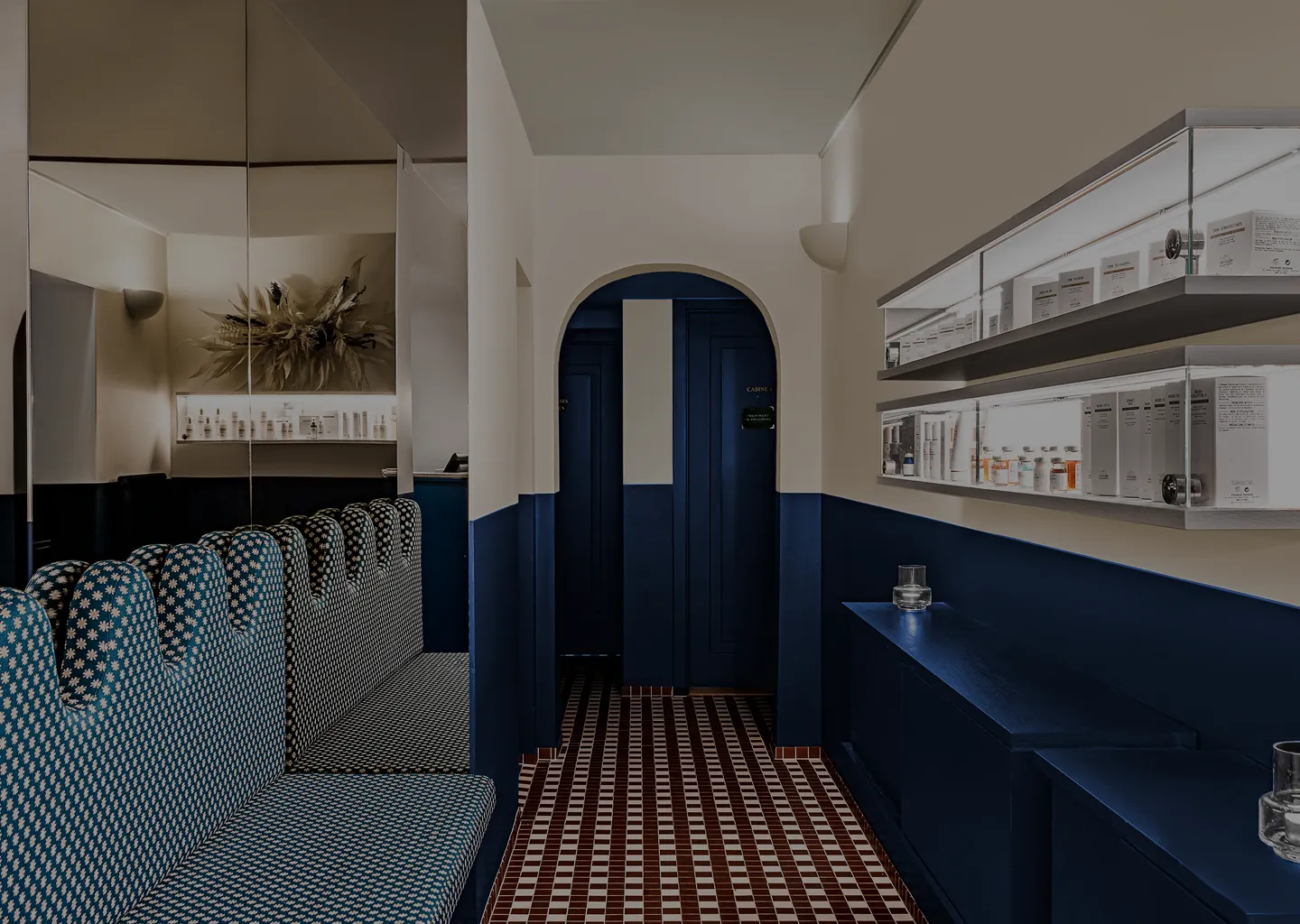 Modern lounge area with patterned blue bench seating, wall-mounted shelves displaying skincare products, and a blue and white tiled floor leading to an arched doorway with dark blue doors.