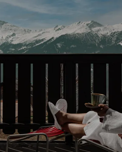 Person in white robe and slippers relaxing on outdoor lounge chair with tea, overlooking snow-capped mountain range.