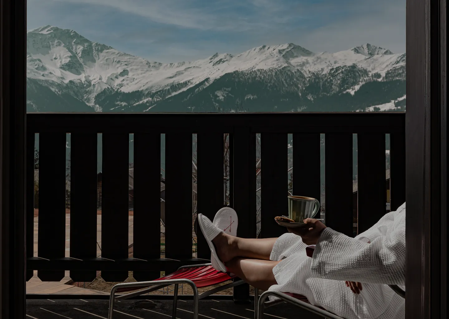 Person in a white bathrobe and slippers sitting on a balcony chair, holding a cup and saucer, overlooking snow-covered mountains.