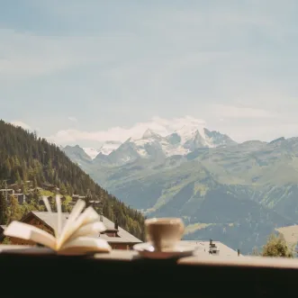Open book and coffee cup on a balcony railing overlooking a scenic mountain landscape with green hills and snow-capped peaks.