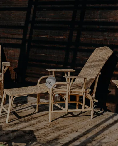 Wicker lounge chair with an ottoman on a wooden deck casting shadows from a slatted overhead structure.