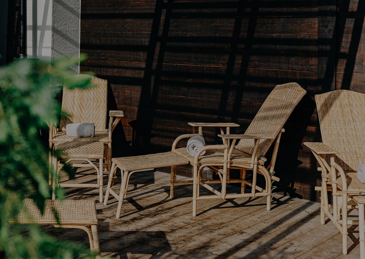 Outdoor wicker lounge chairs and footrests with rolled towels on a wooden deck, casting shadows on a dark wooden wall.