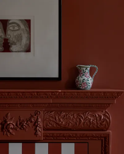 Decorative white pitcher with pink flowers on an intricately carved dark red-brown fireplace mantel beneath a framed abstract face artwork.