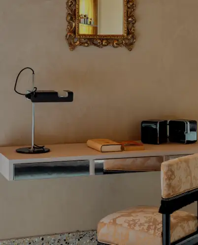Minimalist wall-mounted desk with a modern black lamp, two small black storage boxes, an open book, and a patterned chair below, with a decorative gold-framed mirror on the wall.