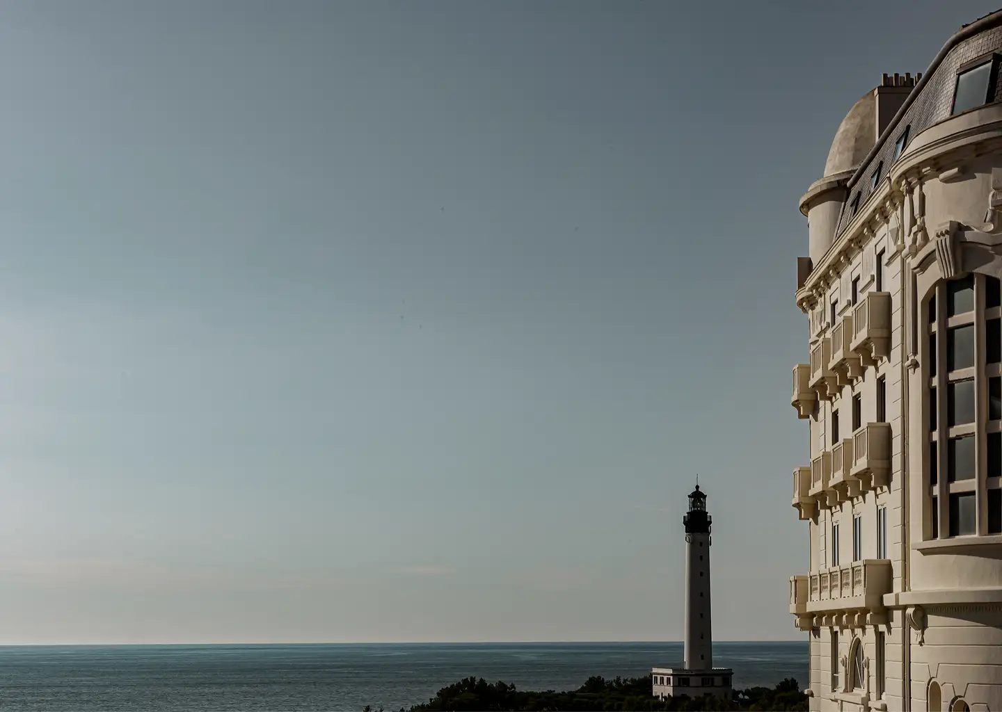 View of a coastal lighthouse near green trees with a large elegant building on the right and calm sea under a clear sky.