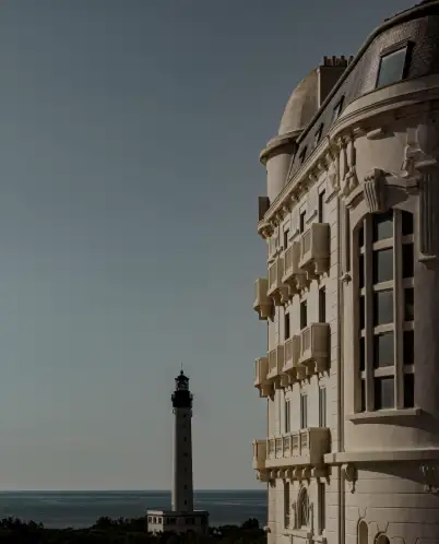 Side view of a historic building with architectural details against a clear sky, with a lighthouse and the ocean in the background.