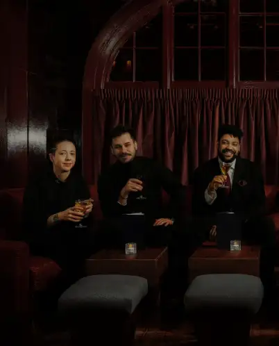 Three people dressed in black sitting on a red couch, holding cocktails, in a dimly lit lounge with a dark red curtain in the background.
