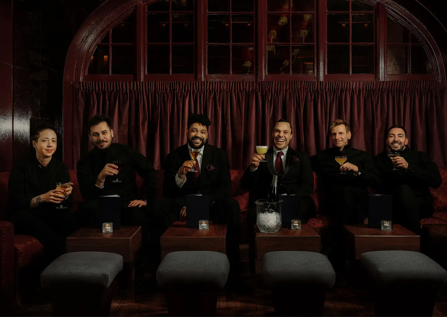 Six people sitting on a red couch in a dimly lit lounge, each holding a cocktail glass and smiling.