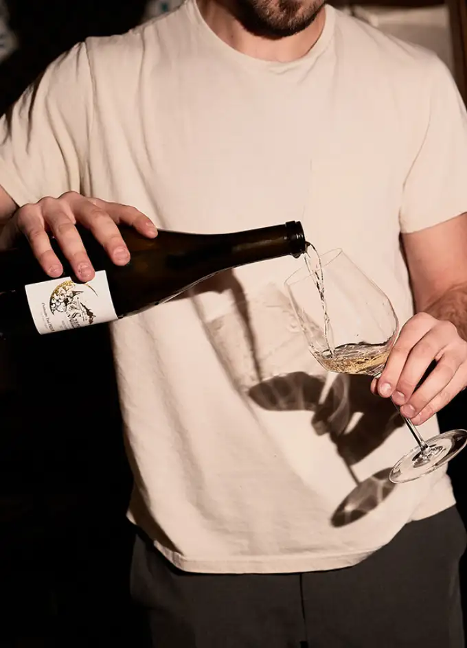 Person wearing a light beige shirt pouring white wine from a dark bottle into a wine glass.