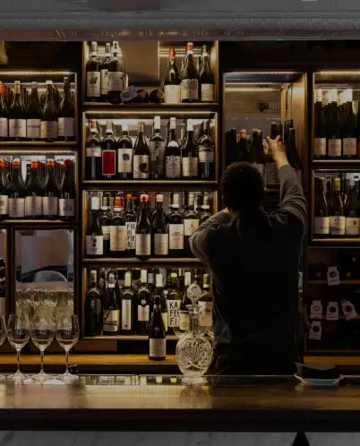 Person selecting a wine bottle from a shelf filled with various wine bottles in a dimly lit bar.