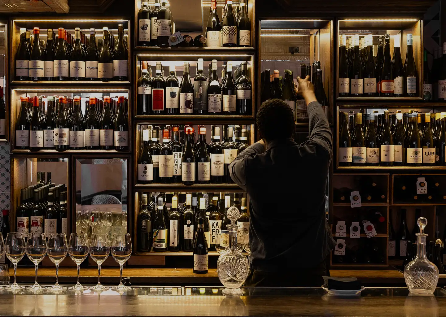 Person reaching for a bottle on illuminated shelves stocked with various wine bottles behind a bar with empty wine glasses and crystal decanters.