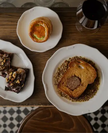 Top-down view of a wooden table with three white plates containing gourmet dishes and a glass of red wine, including toasted bread with a meat filling and lentils.