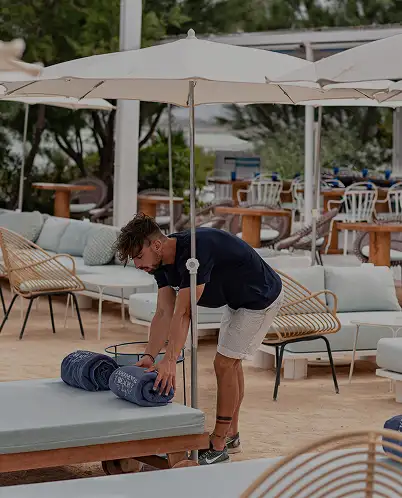 Man in a dark shirt and light shorts rolling up towels on a cushioned outdoor lounge chair under a large white umbrella.