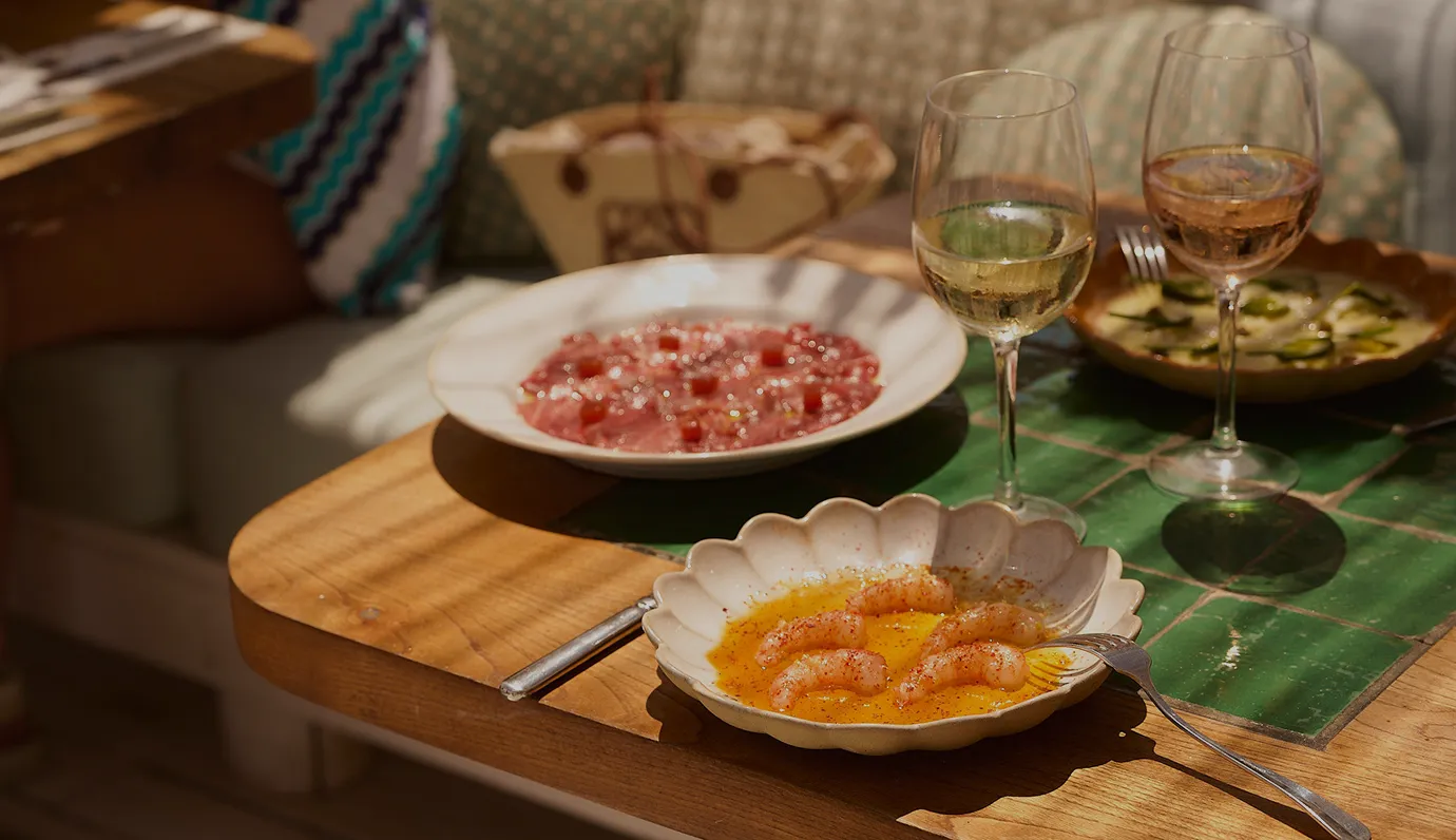 Table with three dishes including shrimp in sauce, a plate of thinly sliced meat, and another dish, accompanied by two glasses of white and rosé wine.