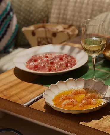 Table with a white wine glass, a plate of shrimp in sauce, and a plate of diced raw fish, possibly sashimi, on a wooden surface.