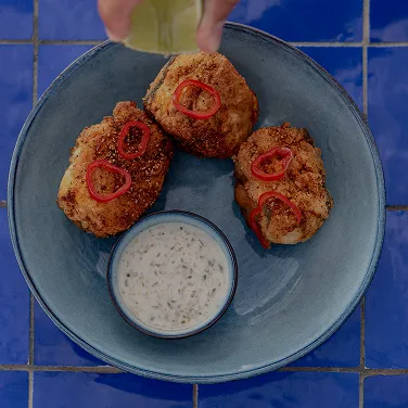 Three breaded fried chicken pieces garnished with red pepper slices on a blue plate with a small bowl of herb dipping sauce, with a hand squeezing lime over the food.