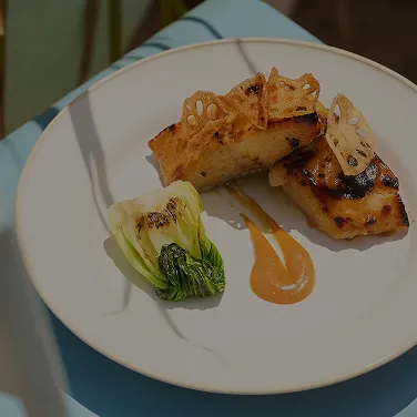 Plate with grilled fish garnished with lotus root chips, a swirl of sauce, and a piece of grilled bok choy.