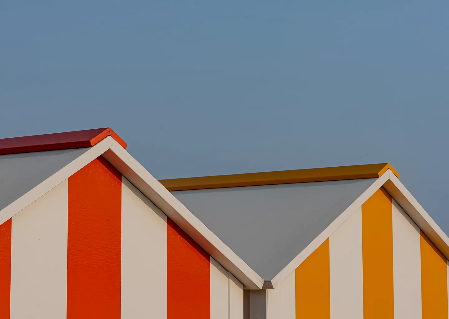 Vista aproximada de duas cabanas de praia com listras verticais, uma em vermelho e branco e outra em amarelo e branco, contra um céu azul claro.