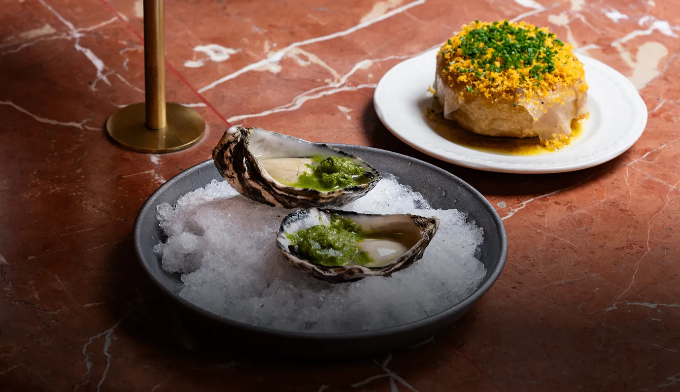Two raw oysters on ice topped with green sauce, with a savory pastry garnished with herbs on a white plate in the background.