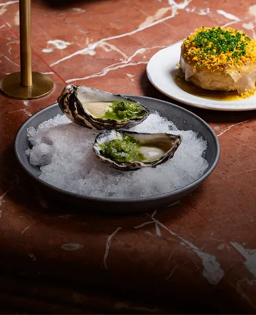 Two oysters on a bed of crushed ice in a gray bowl topped with green garnish, with a round dish covered with yellow topping and herbs in the background on a brown marble surface.