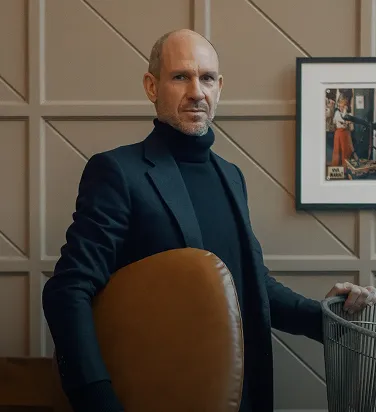 Bald man wearing a black suit and turtleneck sitting on a brown chair in front of a beige patterned wall with a framed picture.