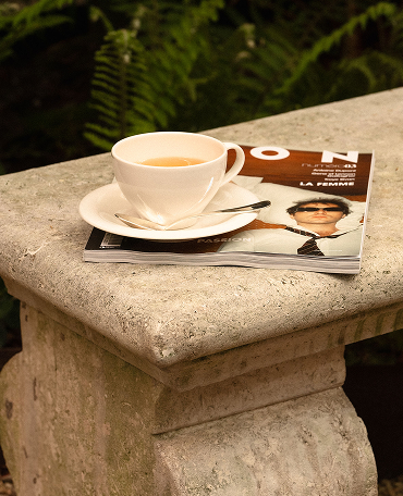 White cup and saucer with a teaspoon resting on a stone bench beside a fashion magazine featuring a man in sunglasses and a tie.