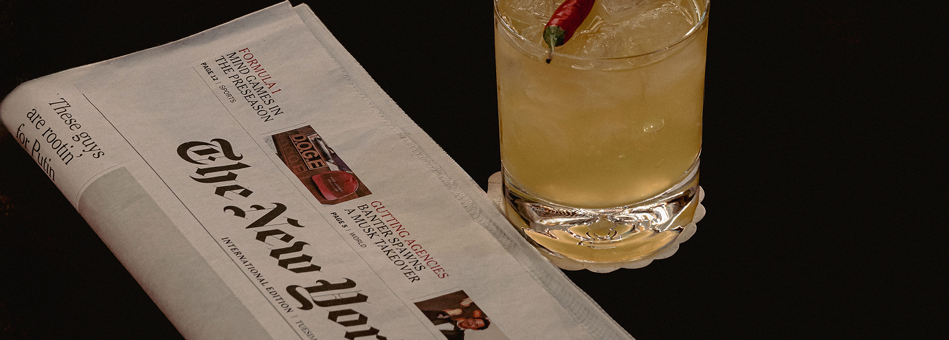 A folded international edition of The New York Times newspaper next to a glass of yellow cocktail garnished with a red chili pepper on a black surface.