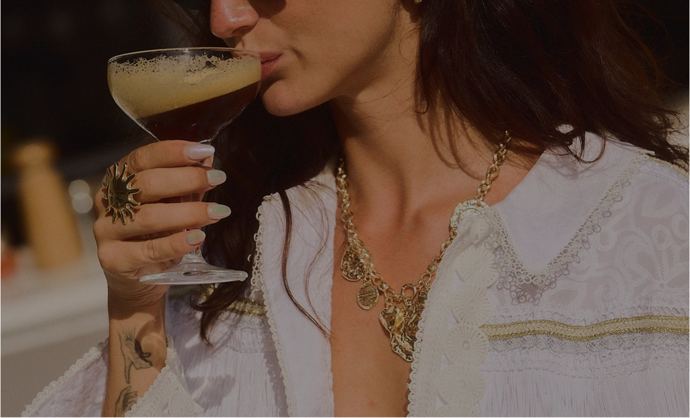 Woman with long hair wearing a white lace garment and gold necklace sipping a dark frothy drink from a coupe glass.