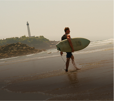 Man walking barefoot on wet beach carrying a surfboard with lighthouse on rocky hill in the background.
