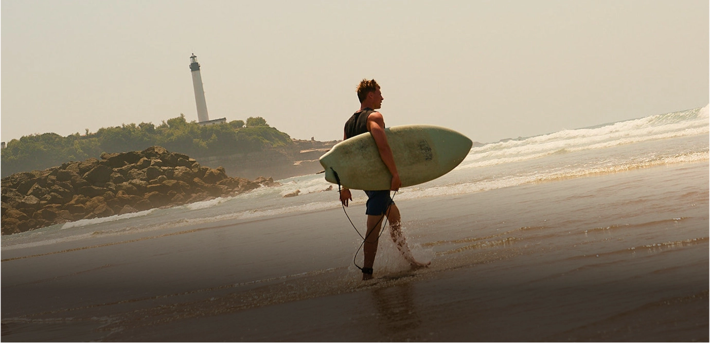 Man carrying a surfboard walking along a sandy beach with waves and a lighthouse on a rocky shore in the background.
