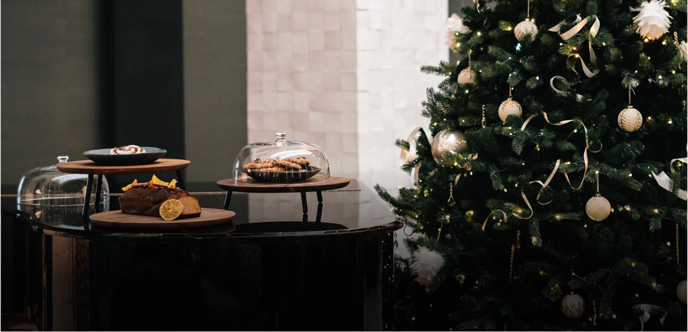 Black piano with wooden stands holding baked goods next to a decorated Christmas tree with lights and white ornaments.
