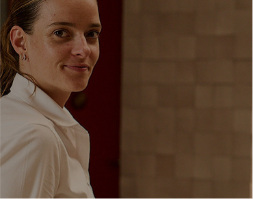 Smiling woman with light skin and brown hair tied back, wearing a white shirt, looking at the camera against a blurred brown background.