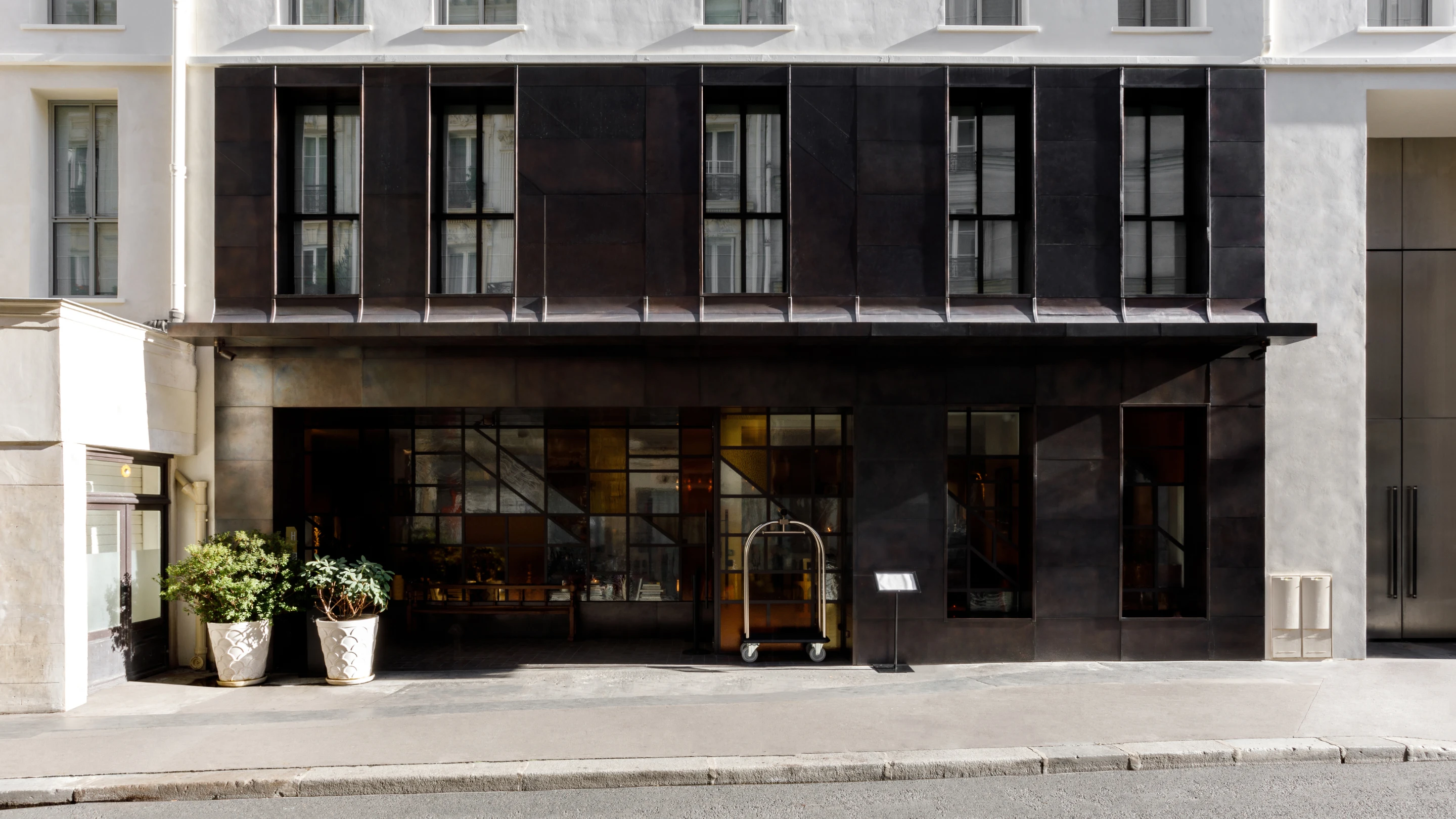 Modern hotel entrance with large dark metal-framed windows, two potted plants, and a luggage cart on the sidewalk.
