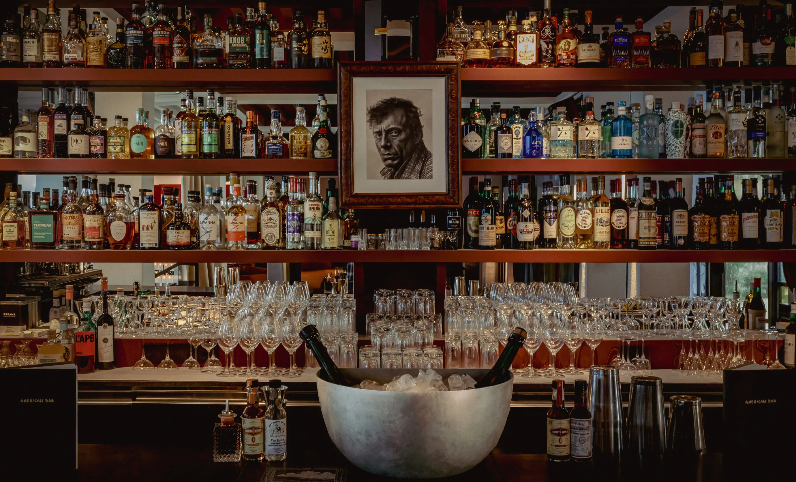 Bar shelves filled with various liquor bottles and glassware, with a large ice bucket holding two champagne bottles in the foreground and a framed black-and-white portrait on the back wall.