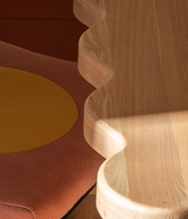 Close-up of a wavy-edged light wooden table surface beside a chair with a brown cushion and a yellow circular patch.