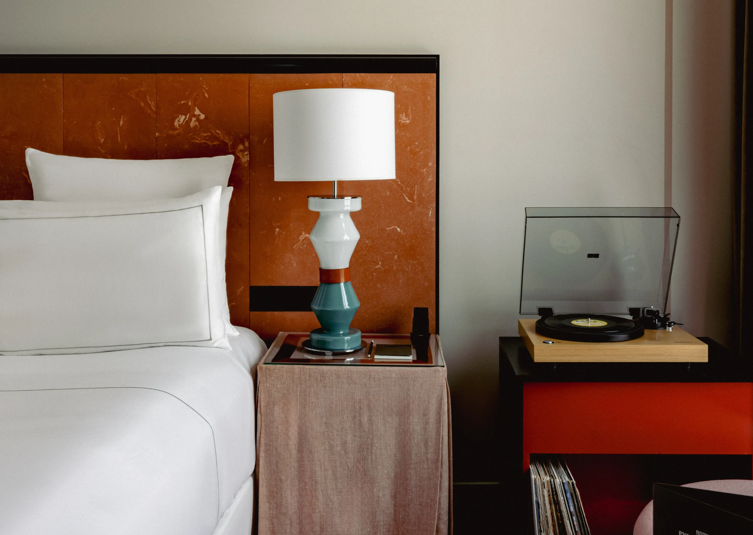 Close-up of a neatly made bed with white linens, a bedside table with a ceramic lamp, and a record player on a black and red cabinet.
