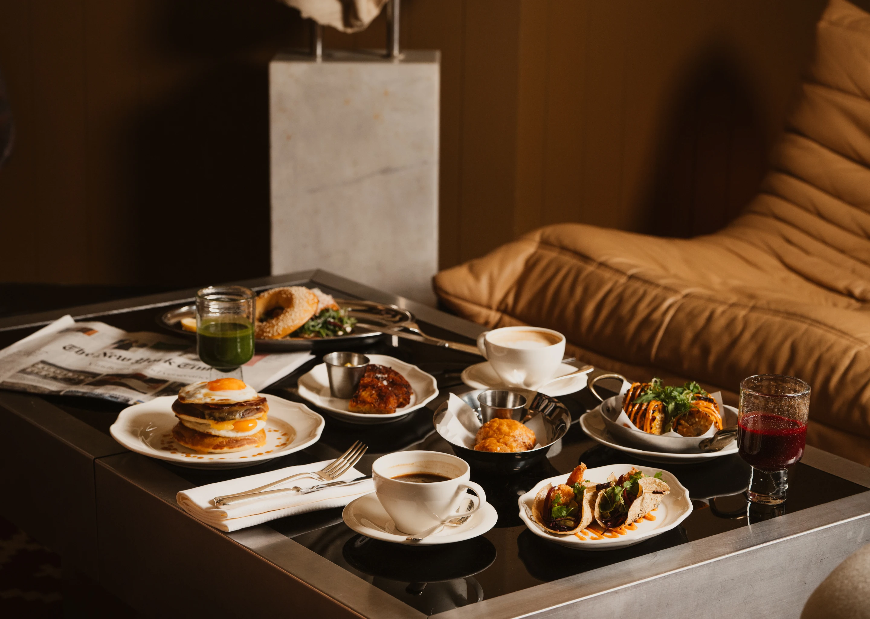 A table with a variety of breakfast foods including pancakes with a fried egg, bagels, tacos, pastries, coffee cups, a green juice, and a glass of red juice in a cozy room with a leather couch.