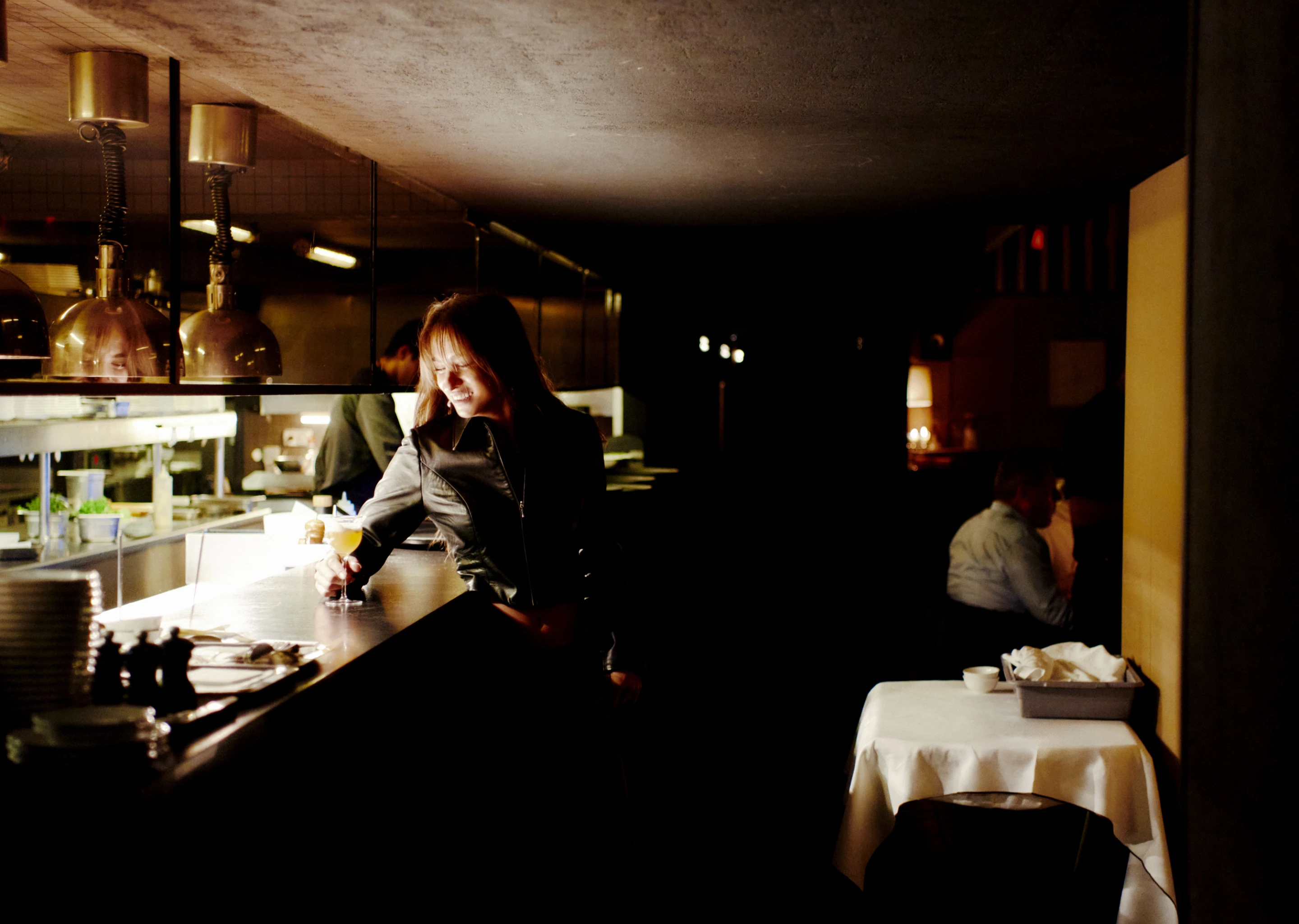 A woman in a black leather jacket smiling and holding a cocktail glass at a dimly lit bar counter.